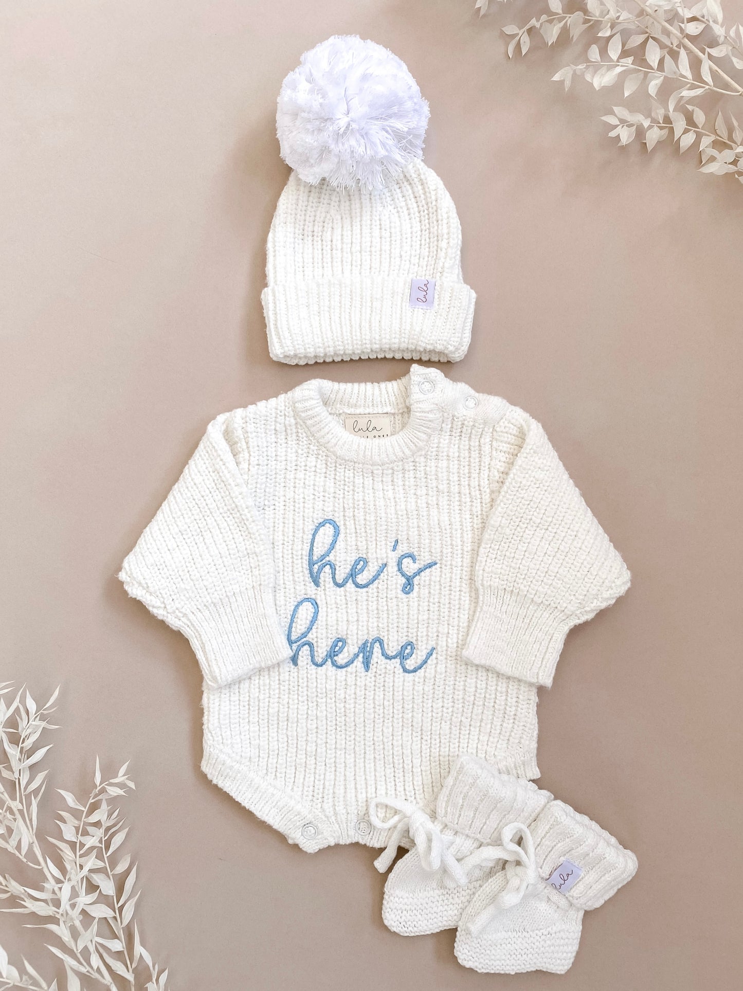 A newborn baby chunky knit ivory coming home outfit with 'he's here' embroidery in blue