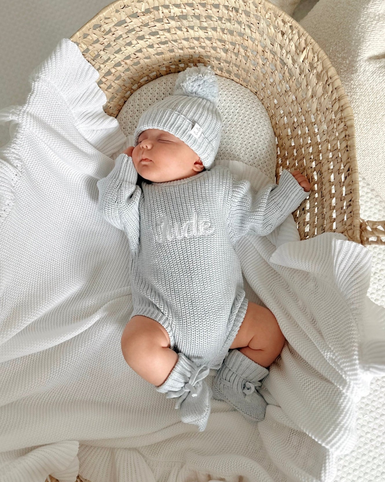 Newborn baby in a blue knit outfit with 'Jude' embroidered on it, lying in a woven baby basket with white blankets.