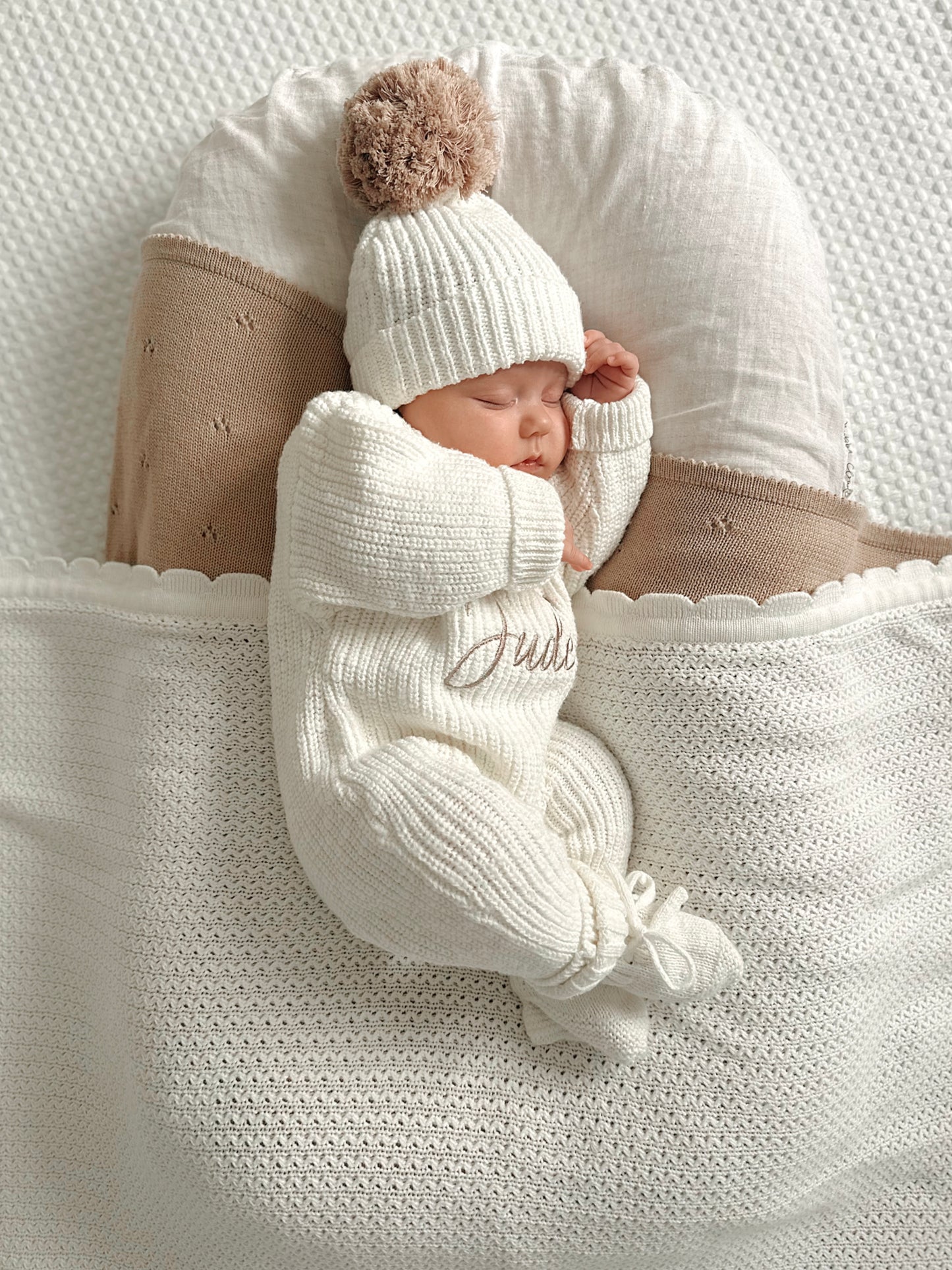 Newborn baby in an ivory knitted romper with his name embroidered on it, wearing a matching hat and booties.