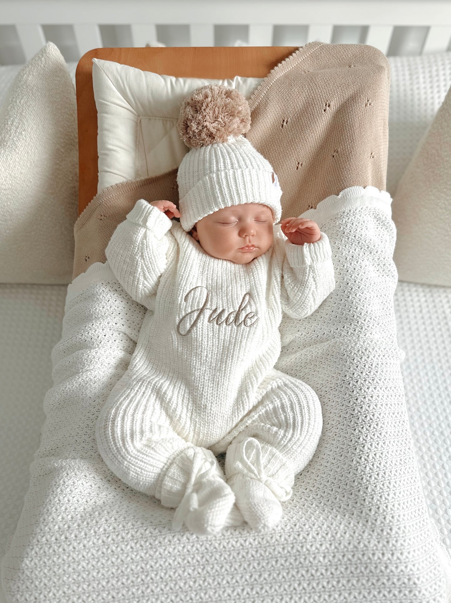 Newborn baby in a white knitted romper with 'Jude' embroidered on it, wearing a matching hat and booties.