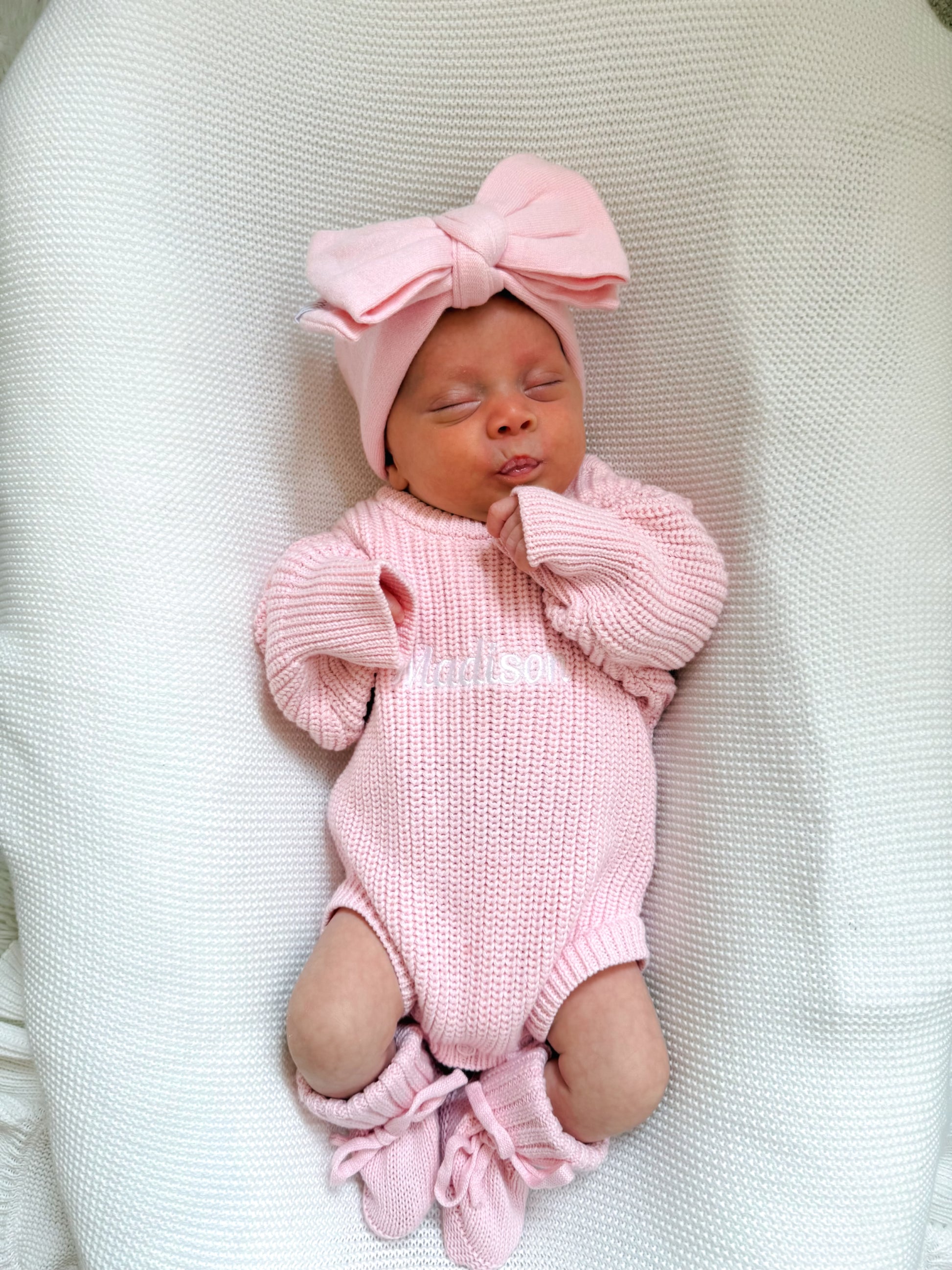 Newborn baby girl in a pink knitted romper with a matching bow on a white blanket