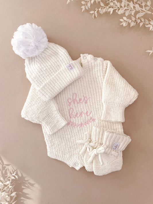 knitted ivory baby outfit set with hat, 'she's here' romper, and booties.