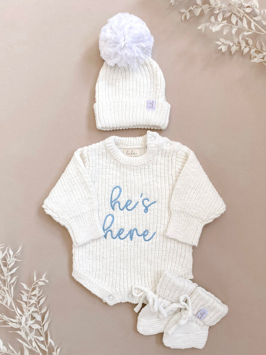 A newborn baby chunky knit ivory coming home outfit with 'he's here' embroidery in blue