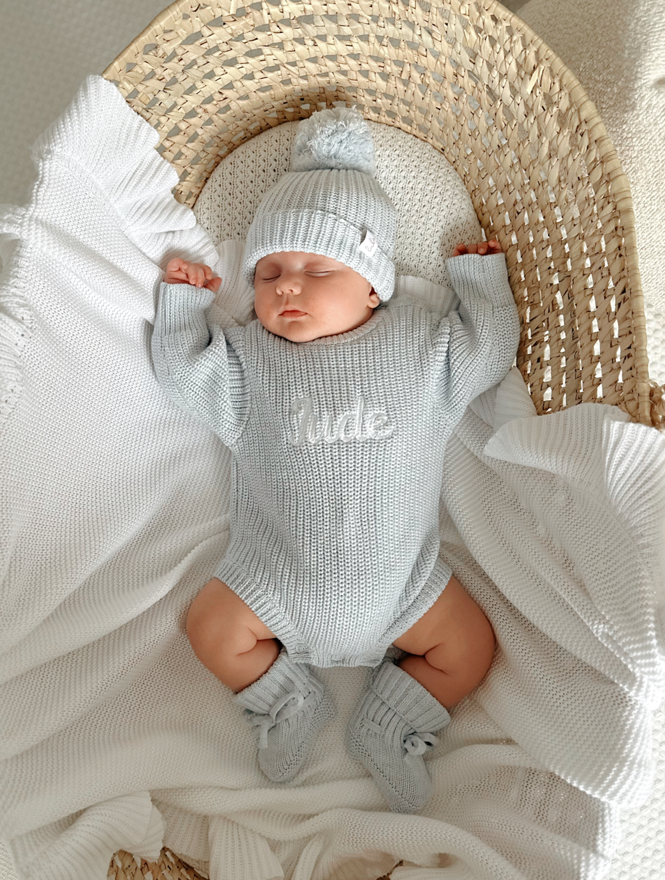Newborn baby in a blue knitted romper, hat and booties lying on a white blanket.