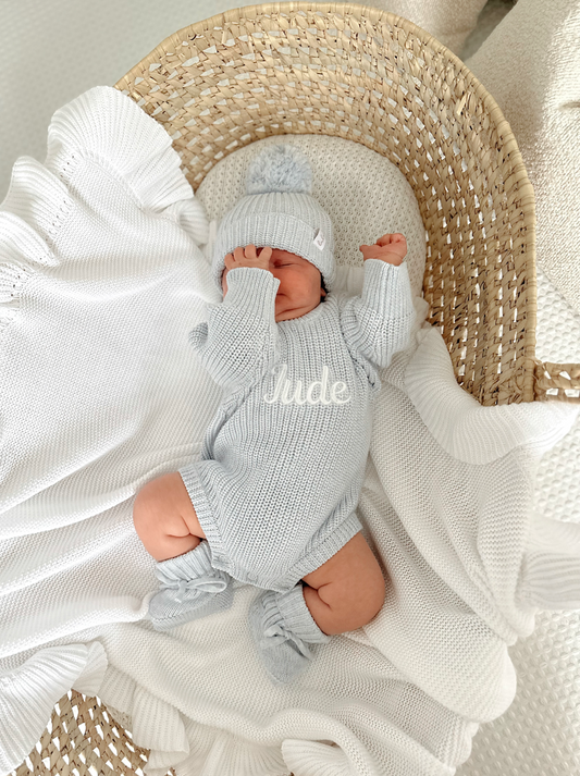 Newborn baby in a blue knitted outfit - romper, hat and booties lying on a white blanket.