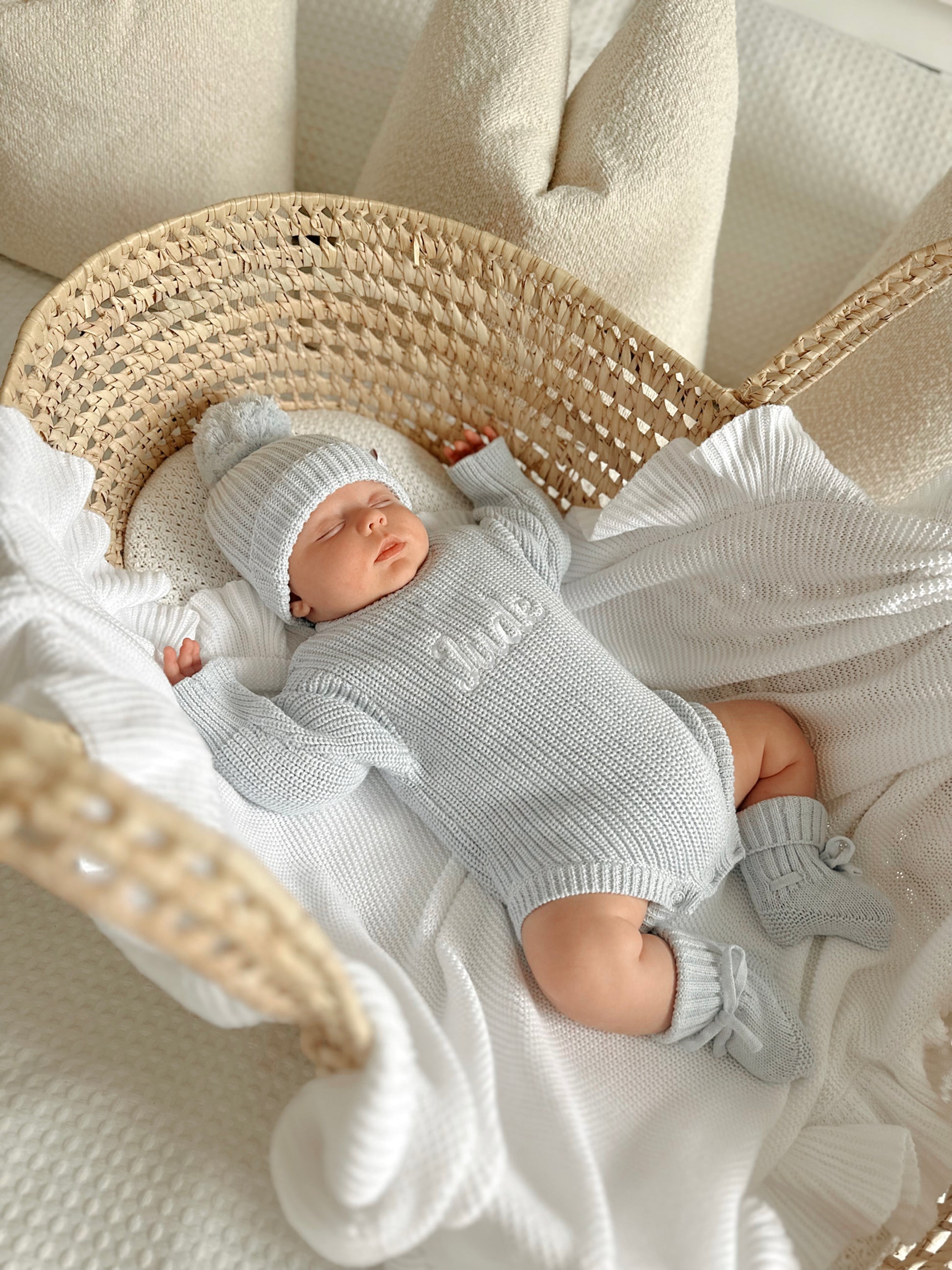 Newborn baby in a blue knitted romper, hat and booties lying on a white blanket.