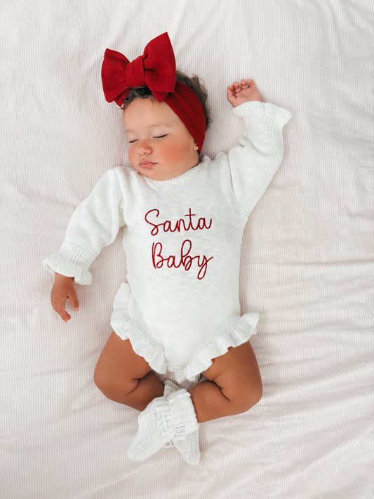 Baby wearing a 'Santa Baby' knitted romper with a red bow on a white background