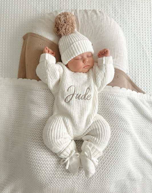 Newborn baby in a white knitted romper with 'Jude' embroidered on it, wearing a matching hat and booties.