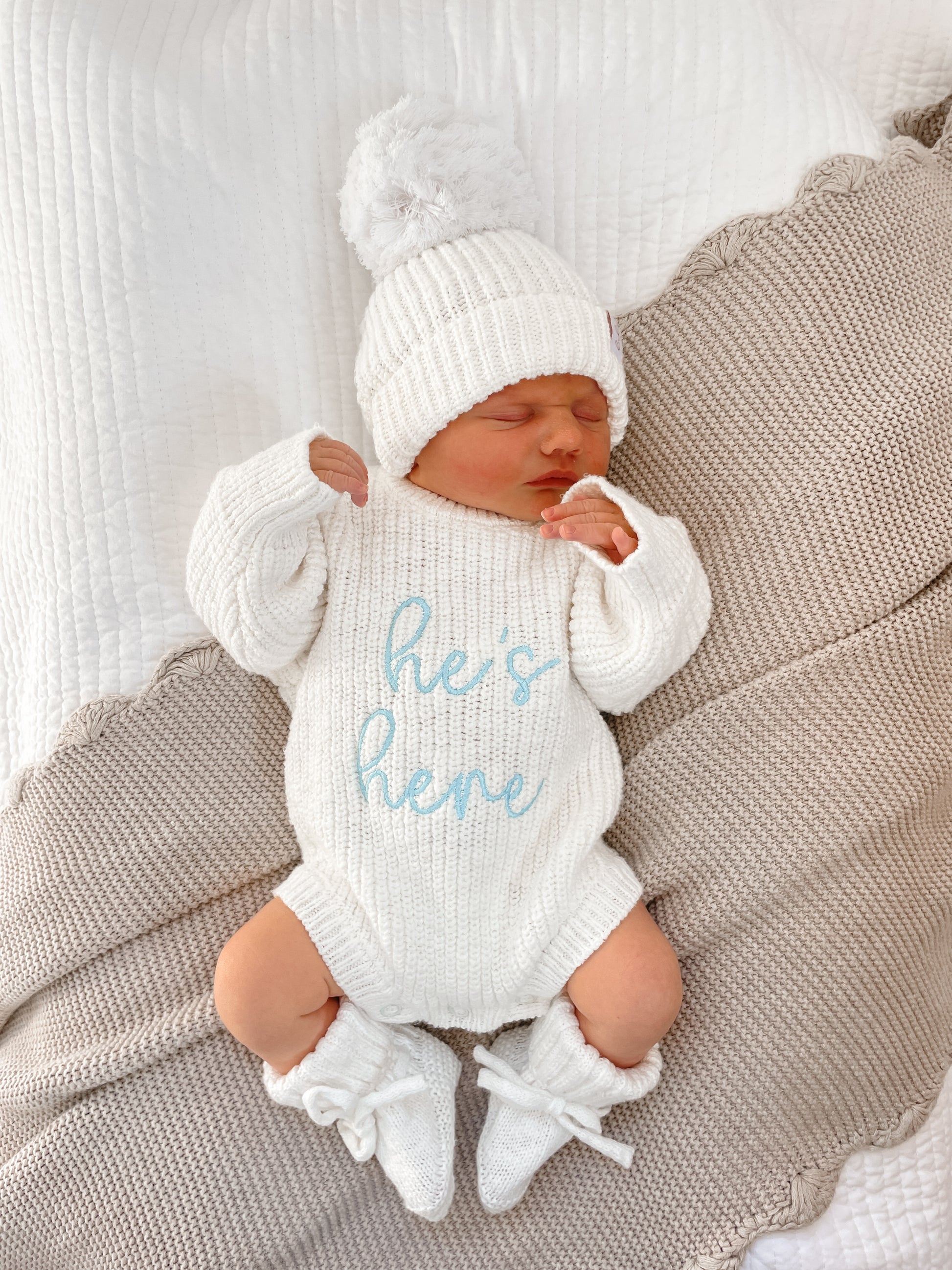 Newborn baby in an ivory outfit with 'he's here' embroidered on it.