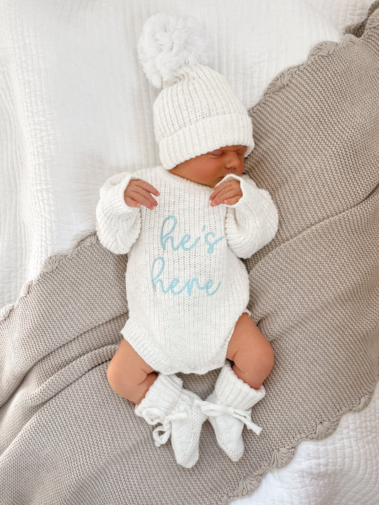 A newborn baby wearing a chunky knit ivory coming home outfit with 'he's here' embroidery in blue.