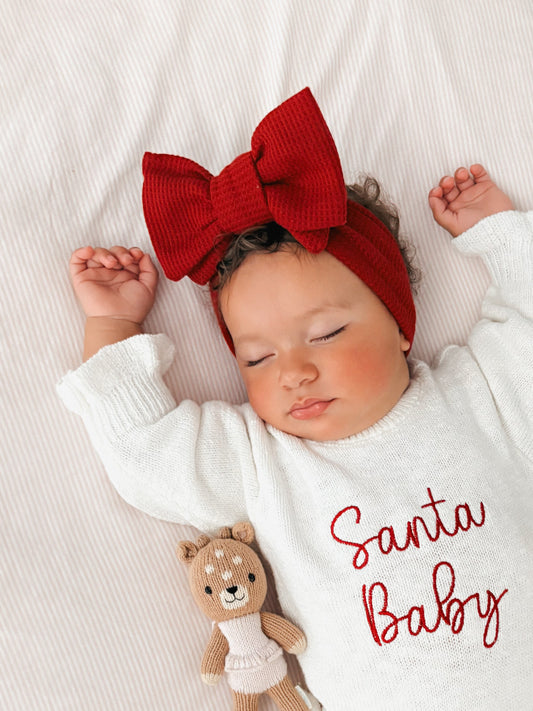 baby wearing a 'Santa Baby' Christmas outfit with a red bow on a white blanket.