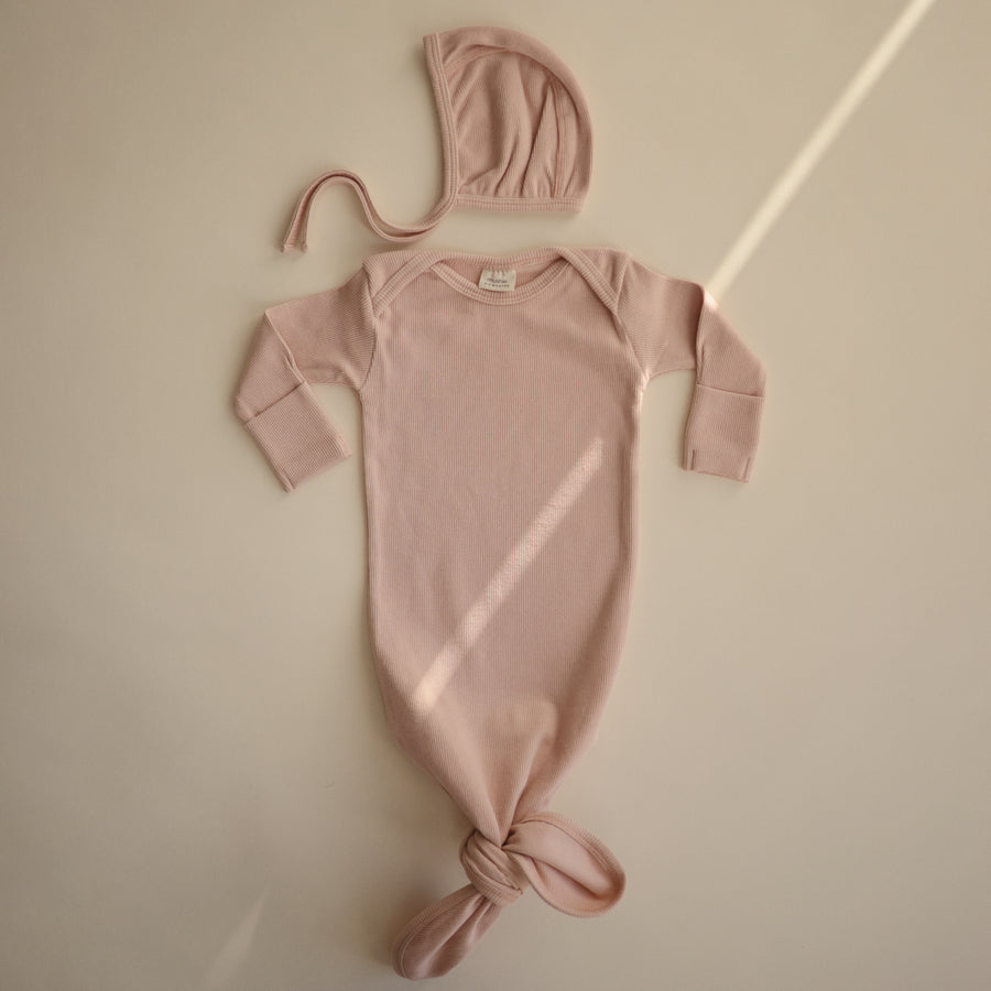 Flat lay of a Mushie ribbed knotted baby gown in blush with matching bonnet 